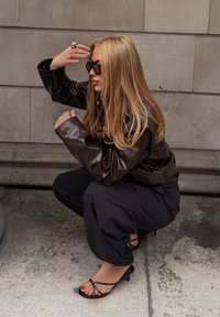 Zwarte leren jas, losse zwarte broek en zwarte sandalen met banden. Lang blond haar met gemanicuurde nagels, geposeerd tegen een stenen muur.