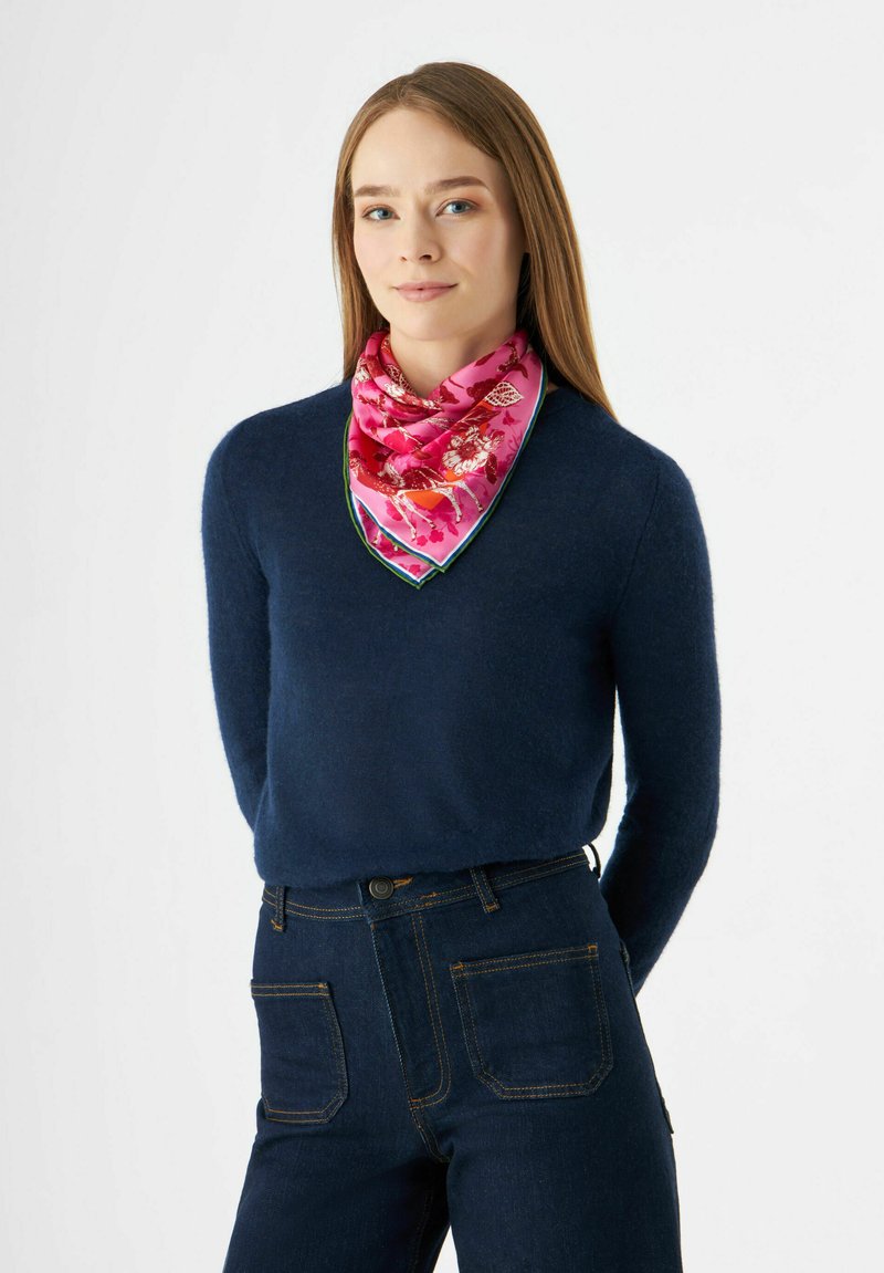 Écharpe en soie rose avec un motif floral et une bordure verte, nouée autour du cou, portée avec un pull marine et un jean foncé. Texture lisse.