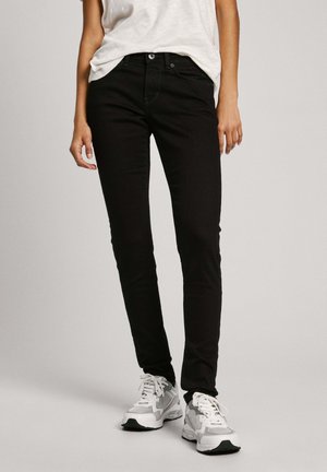 Personne portant un jean slim noir, des baskets blanches décontractées et un haut sans manches blanc rentré dans le pantalon, debout sur un fond neutre.