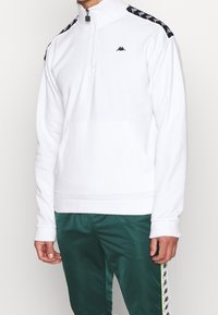 Bílý pulovr s půlvložkou na zip vyrobený z měkké látky, s černým logem Kappa a pruhy s motivem pepita podél ramen. Součástí je přední kapsa.