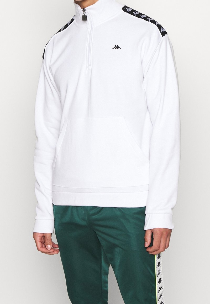 Bílý pulovr s půlvložkou na zip vyrobený z měkké látky, s černým logem Kappa a pruhy s motivem pepita podél ramen. Součástí je přední kapsa.