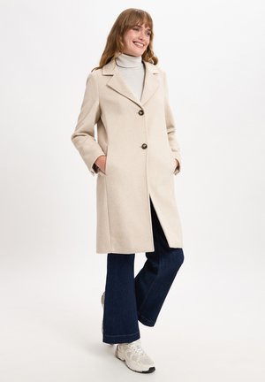 Beige Wollmischungmantel mit einem Schalkragen, zwei Fronttaschen und zwei dunklen Knöpfen. Getragen über einem hellen Rollkragenpullover und flared dunklen Jeans.