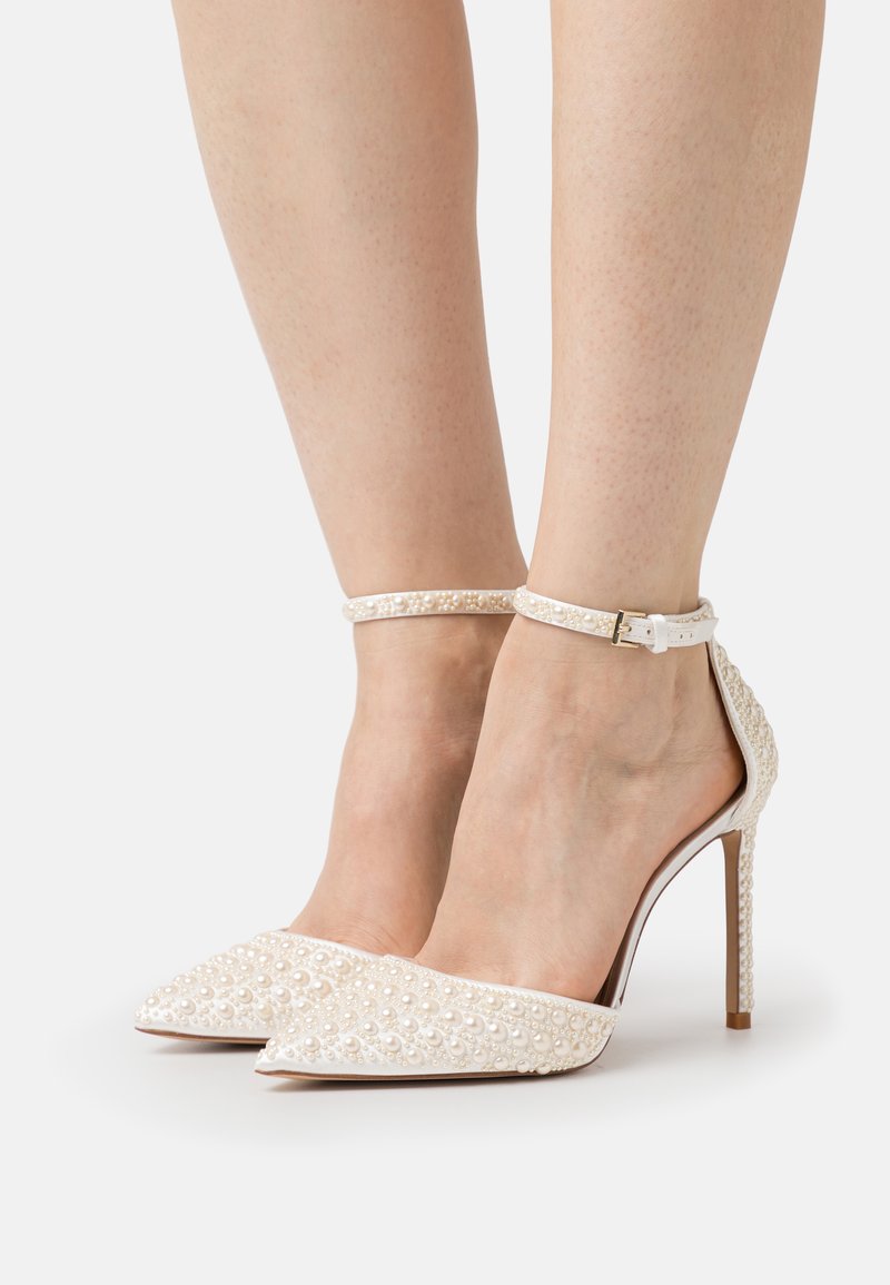 Scarpe bianche con tacco alto e punta affusolata, con un motivo testurizzato di piccole perle e una delicata cinghia alla caviglia con una fibbia metallica.