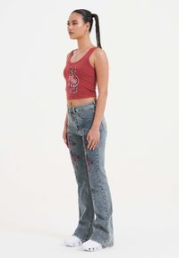 Top corto sin mangas rojo con un diseño gráfico, combinado con jeans acampanados de color azul claro con bordado floral y zapatillas blancas.