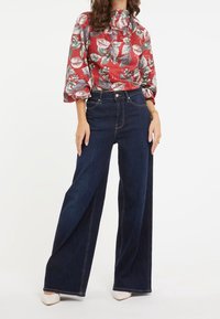 Rode bloes met bloemenprint en pofmouwen; hoge taille, wijde blauwe denim jeans; gecombineerd met witte hakken.
