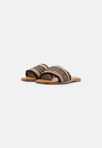 Bruna slip-on sandaler med två breda remmar som har svart frans och ett geometriskt mönster. Sullan är platt och gjord av ljust trä.