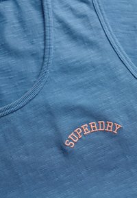 Niebieski sportowy top bez rękawów z teksturowanej tkaniny, z okrągłym dekoltem i wyraźnym logo "SUPERDRY" w różowym haftowanym napisie.