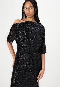 Abito nero di paillettes con scollatura a spalle scoperte, maniche a cappuccio e silhouette aderente. La texture presenta righe di paillettes verticali per un effetto scintillante.