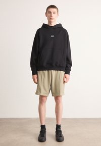 Black hooded sweatshirt with a front pocket and small logo, paired with beige fleece shorts. Black sneakers and dark socks complete the look.