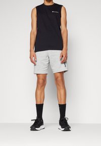 Svart ärmlös topp med logotyp, grå shorts, svarta strumpor och svarta träningsskor. Modellen står mot en enfärgad bakgrund.