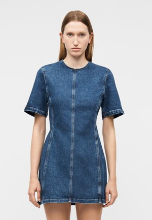 Kobieta w dopasowanej, krótkorękawowej niebieskiej sukience jeansowej z detalami przeszyć z przodu, stojąca na tle jednolitego jasnoszarego tła.