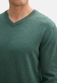 Pulover verde cu decolteu în V fabricat dintr-un material moale, texturat. Prezintă un logo discret pe piept și detalii ribbed la decolteu și la mansete.