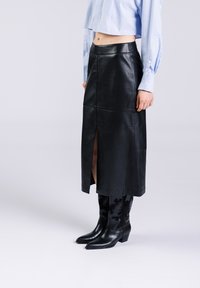 Zwart leren midi-rok met een split aan de voorkant, met een gladde textuur. Gecombineerd met zwarte cowboylaarzen. Lichtblauw crop shirt zichtbaar erboven.