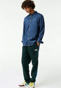 Giovane che indossa una camicia di denim con bottoni, pantaloni da jogging scuri con un logo e sneaker colorate, in piedi con le mani in tasca e lo sguardo rivolto verso il basso.