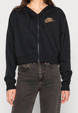 Mujer vestida con una sudadera con cremallera negra y un logo de Nike con estampado de leopardo, y jeans de cintura alta en gris oscuro, de pie con los brazos relajados.