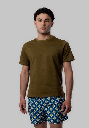 Giovane uomo in piedi che indossa una camicia verde oliva a maniche corte e pantaloncini blu con motivo geometrico su sfondo grigio.