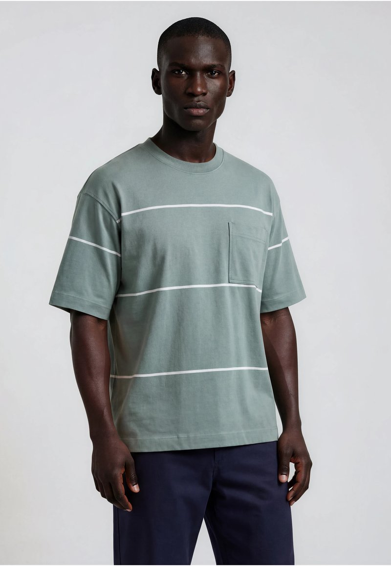 Homme portant un T-shirt vert atténué à manches courtes avec des rayures horizontales blanches et une poche poitrine, associé à un pantalon bleu marine foncé.