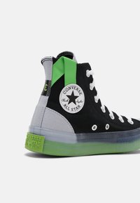 Zapatilla alta con parte superior de lona negra, acentos verdes, secciones de tela gris, cordones blancos y una suela de goma verde translúcida.
