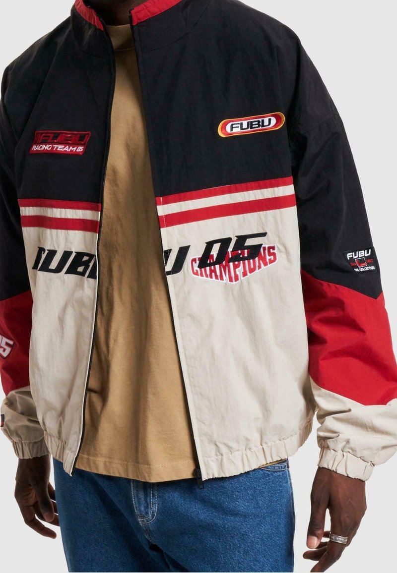 Veste bicolore en bleu marine, rouge et beige. Présente la marque FUBU, un col montant et des poignets élastiques. Texture lisse avec fermeture éclair.