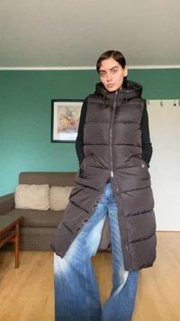 Czarna długa, puchowa kamizelka z kapturem, z przodu na zamek i z kieszeniami po bokach, noszona na niebieskich szerokich spodniach dżinsowych. Miękka, pikowana faktura.