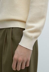 Maglione beige chiaro testurizzato con orlo e polsini a costine, abbinato a pantaloni plissettati verde oliva, che mostrano un tessuto morbido e motivi dettagliati.