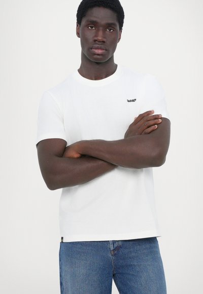 T-shirt en coton blanc avec un col rond et des manches courtes. Présente un petit logo noir sur la poitrine. Associé à un jean en denim bleu.