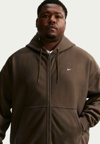 Hombre con sudadera marrón con cremallera y pequeño logo blanco de Nike, de pie con las manos en los bolsillos contra un fondo claro y sencillo.