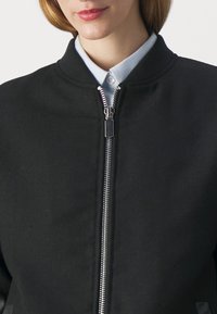 Femme portant une veste noire zippée par-dessus une chemise blanche à col, montrant la partie supérieure de la poitrine et le cou.