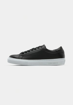 Black leather sneaker with a smooth finish, white rubber sole, flat laces, and a minimalistic design. Features embossed branding on side.