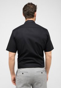Zwart overhemd met korte mouwen en een gladde textuur, gedragen met lichtgrijze gepaste broek met twee achterzakken.
