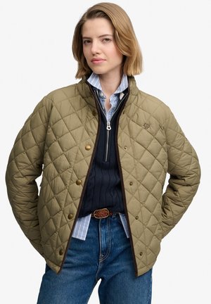 Mujer joven con una chaqueta acolchada color oliva sobre un jersey azul marino con media cremallera, camisa a rayas, cinturón trenzado y vaqueros azules con las manos en los bolsillos.