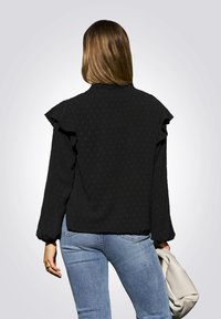 Zwarte blouse met lange mouwen, ruches op de schouders, een textuurpatroon en een rechte zoom. Draagt het met blauwe jeans en houdt een lichtgekleurde handtas vast.