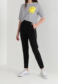 Stribet sort og hvid t-shirt med en gul smiley face lomme, parret med sorte tapered bukser og hvide sneakers.