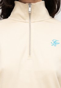Μπεζ φλις με φερμουάρ και ψηλό γιακά, με ασημένιο φερμουάρ και ανοιχτό μπλε κεντημένο λογότυπο. Μαλακή υφή, άνετη εφαρμογή.