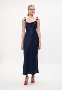 Rochie din satin albastru marin, cu decolteu tip cowl și accente florale pe bretele. Silueta ajustată, care se îngustează într-o lungime midi. Asortată cu sandale roșii cu barete.