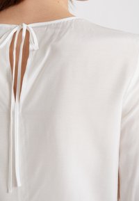 Vit blus med öppen rygg som säkras med snörningar, mjuk textur och avslappnad passform. Accentuerad med en subtil lyster.