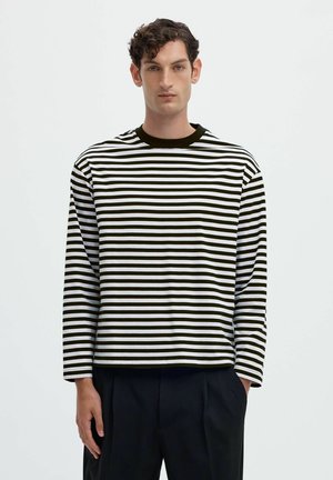 Jeune homme aux cheveux courts et bouclés portant un t-shirt à manches longues rayé horizontalement noir et blanc et un pantalon noir, debout avec une main dans la poche.