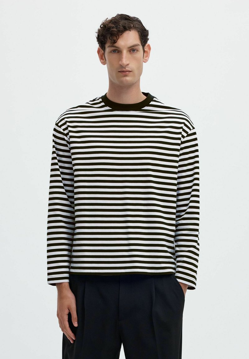 Jeune homme aux cheveux courts et bouclés portant un t-shirt à manches longues rayé horizontalement noir et blanc et un pantalon noir, debout avec une main dans la poche.