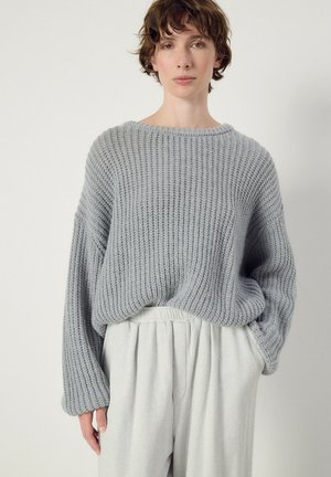 Personne aux cheveux courts, bouclés et bruns, portant un pull gris côtelé ample et un pantalon taille haute gris clair, debout devant un fond uni.