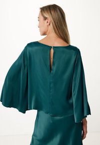 Blusa di seta teal con maniche a campana e apertura a chiave sul retro, caratterizzata da una texture liscia e un design fluido.
