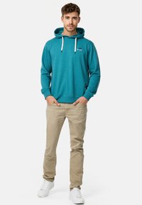 Teal hættetrøje med snoretræk, ribbede manchetter og et lille logo. Paarret med beige bukser og hvide sneakers, stående på en hvid baggrund.