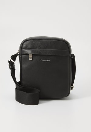 Sac en cuir noir à bandoulière avec une surface texturée, fermeture éclair et sangle réglable. Il dispose d'une poche avant et d'une marque en argent.