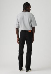 Muškarac s pletenom kosom nosi široku sivu majicu, crne traperice s kožnim detaljem i crne tenisice, stoji okrenut leđima.