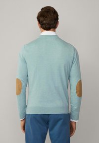 Maglione azzurro chiaro realizzato in materiale morbido con scollatura a V, dotato di toppe beige sui gomiti e polsini a coste. Indossato sopra una camicia bianca.