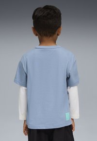 Vaikas esančių nugara, dėvintis šviesiai mėlyną trumpasluoksnį marškinį ant baltų ilgų rankovių, su juodomis kelnėmis prieš pilką foną.