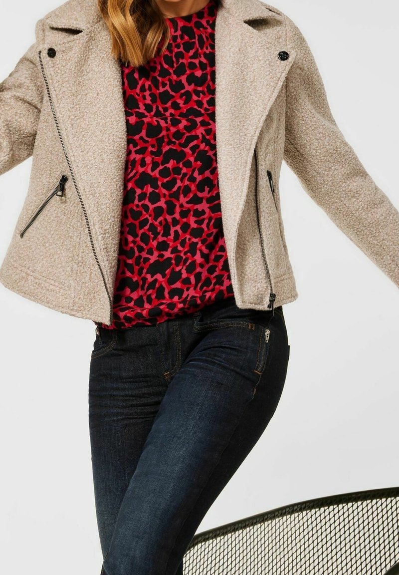 Béžová motorkářská bunda z vlněné směsi s texturovanou tkaninou, asymetrickým zipem a dvěma bočními kapsami, navrstvená přes top s červeným a černým leopardím potiskem.