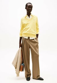 Geel V-hals trui over een wit overhemd met kraag, gecombineerd met beige wijde broek en zwarte loafers. Draagt een beige jas en handtas.