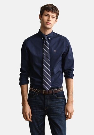 Jeune homme portant une chemise bleu foncé boutonnée avec des manches retroussées, une cravate rayée bleu marine, une ceinture à motifs et un jean foncé sur un fond uni.