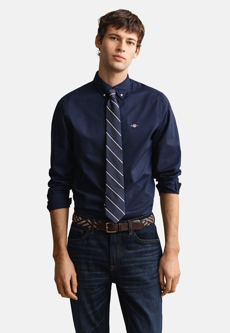 Jeune homme portant une chemise bleu foncé boutonnée avec des manches retroussées, une cravate rayée bleu marine, une ceinture à motifs et un jean foncé sur un fond uni.