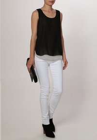 Top nero senza maniche con un design fluttuante, sovrapposto a una base bianca, abbinato a jeans skinny bianchi e stivaletti neri.